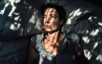 A woman lies on a bed, looking upward with a thoughtful expression. Dappled sunlight and shadows from leaves create patterns across her face, pillow, and shirt.