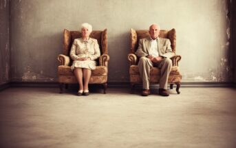 An elderly woman and man sit separately in vintage armchairs against a worn, gray wall, both dressed formally and looking straight ahead. The setting appears quiet and sparse, with empty space around them.